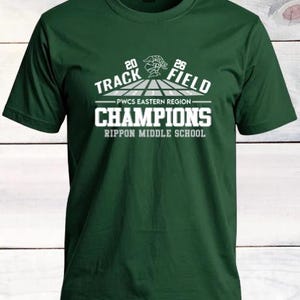 May include: A dark green t-shirt with a white graphic design. The design includes the words "Track Field Champions" with the years "20" and "26" and the text "PWCS Eastern Region" and "Rippon Middle School".