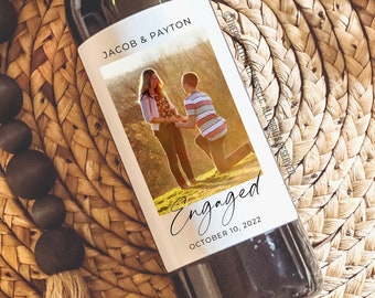 May include: A 4x5 inch wine bottle label with a photo of a couple, the man is on one knee, and the text "Engaged" with the date "October 10, 2022".