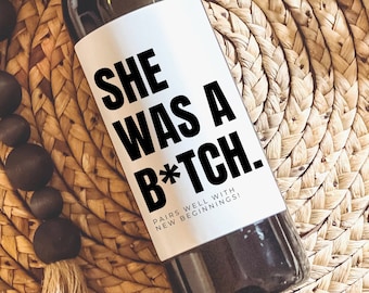 Może przedstawiać: Butelka wina z białą etykietą z napisem "SHE WAS A B*TCH." czarnymi literami. Etykieta zawiera również tekst "PAIRS WELL WITH NEW BEGINNINGS!". Butelka stoi na tkanej powierzchni.