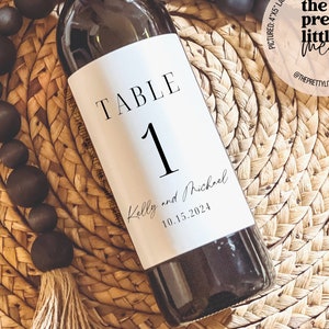 May include: A white wine bottle label with black text that reads "TABLE 1 Kelly and Michael 10.15.2024". The label is on a bottle of red wine. The bottle is sitting on a woven straw mat.