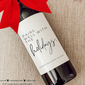 May include: A white wine bottle label with black text that reads "Pairs well with the holidays."  The label is on a bottle of red wine with a red bow on the neck. The text "Wishing you a happy holiday." is at the bottom of the label.