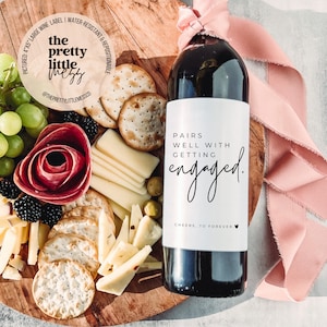 Puede incluir: Una etiqueta de botella de vino blanco con texto negro que dice "Pairs well with getting engaged. Cheers, to forever." La botella est&aacute; sobre una tabla de madera con una cinta rosa y una tabla de queso y galletas.
