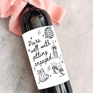 May include: A dark wine bottle with a white label featuring the text "Pairs well with getting engaged." The label includes illustrations of a disco ball, champagne bottle, and glasses. A pink ribbon is tied around the bottle's neck.