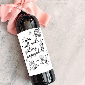 May include: A dark wine bottle with a white label that reads "Pairs well with getting engaged." The bottle is adorned with a light pink ribbon. The label features illustrations of a disco ball, champagne bottle, and champagne glasses.
