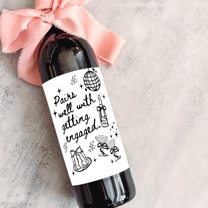 May include: A dark wine bottle with a white label that reads "Pairs well with getting engaged." The bottle is adorned with a light pink ribbon. The label features illustrations of a disco ball, champagne bottle, and champagne glasses.