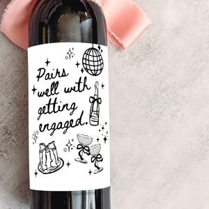 May include: A dark wine bottle with a white label featuring the text "Pairs well with getting engaged." The label includes illustrations of a disco ball, champagne bottle, cake, and champagne glasses. A pink ribbon is in the background.