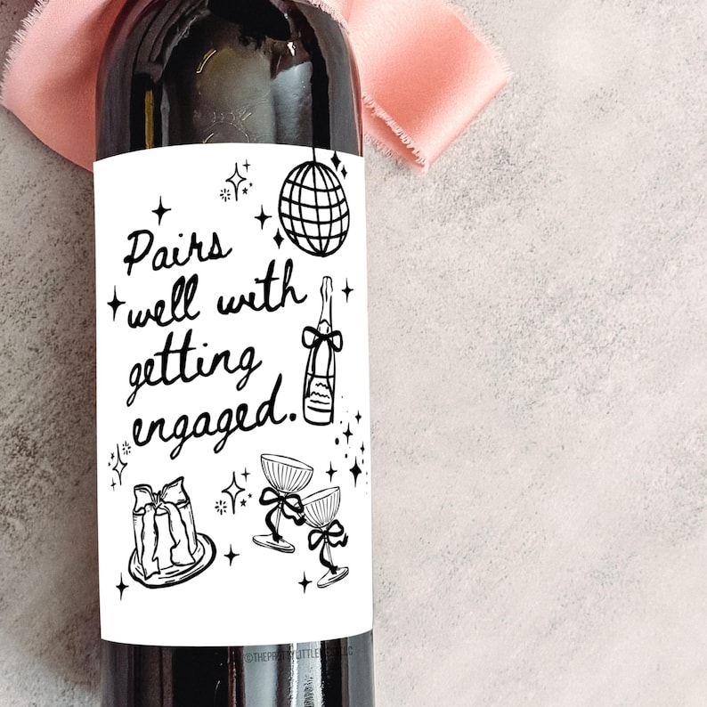 May include: A dark wine bottle with a white label featuring the text "Pairs well with getting engaged." The label includes illustrations of a disco ball, champagne bottle, cake, and champagne glasses. A pink ribbon is in the background.