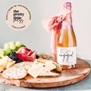 Puede incluir: Una peque&ntilde;a botella de champ&aacute;n con una cinta rosa atada alrededor del cuello. La botella tiene una etiqueta blanca con el texto "Pairs well with getting engaged." La botella est&aacute; sentada sobre una superficie de madera con un plato de queso y galletas.