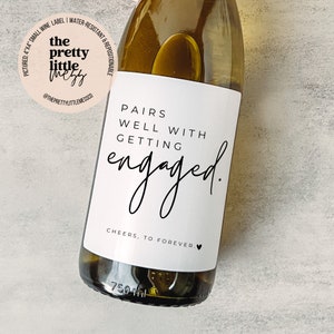 Puede incluir: Una botella de vino blanco con una etiqueta blanca que dice "Pairs well with getting engaged." y "Cheers, to forever." con un peque&ntilde;o s&iacute;mbolo de coraz&oacute;n.