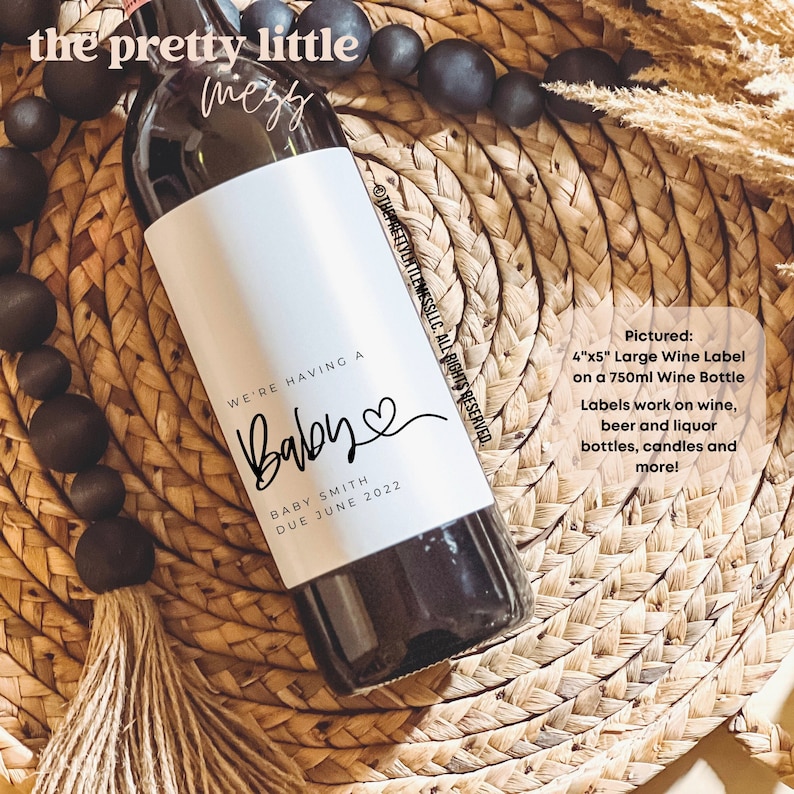 May include: A white wine bottle label with black text that reads "We're having a Baby" with a heart symbol and the text "Baby Smith Due June 2022". The label is on a 750 ml wine bottle. The text "Pictured: 4"x5" Large Wine Label on a 750ml Wine Bottle Labels work on wine, beer and liquor bottles, candles and more!" is in the upper right corner of the image.