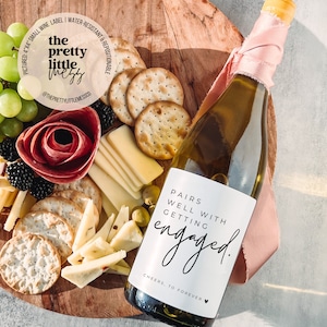 Puede incluir: Una botella de vino blanco con una etiqueta que dice "Pairs well with getting engaged." La botella est&aacute; envuelta en una cinta rosa y est&aacute; sobre una tabla de madera con queso, galletas y uvas.