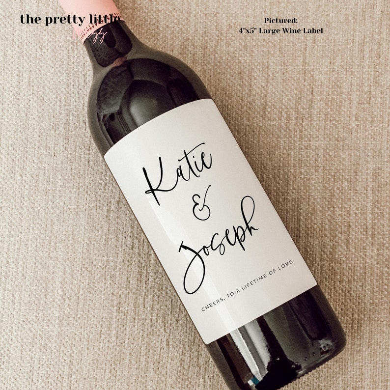 May include: A white wine bottle label with black calligraphy that reads "Katie & Joseph" and "CHEERS, TO A LIFETIME OF LOVE."