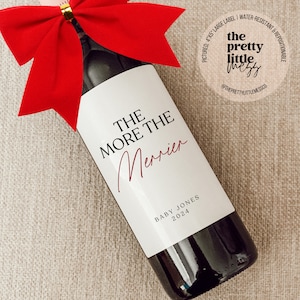 May include: A white wine bottle label with the text "THE MORE THE MERRIER" in black and red lettering. The label also includes the text "BABY JONES 2024" in black lettering.