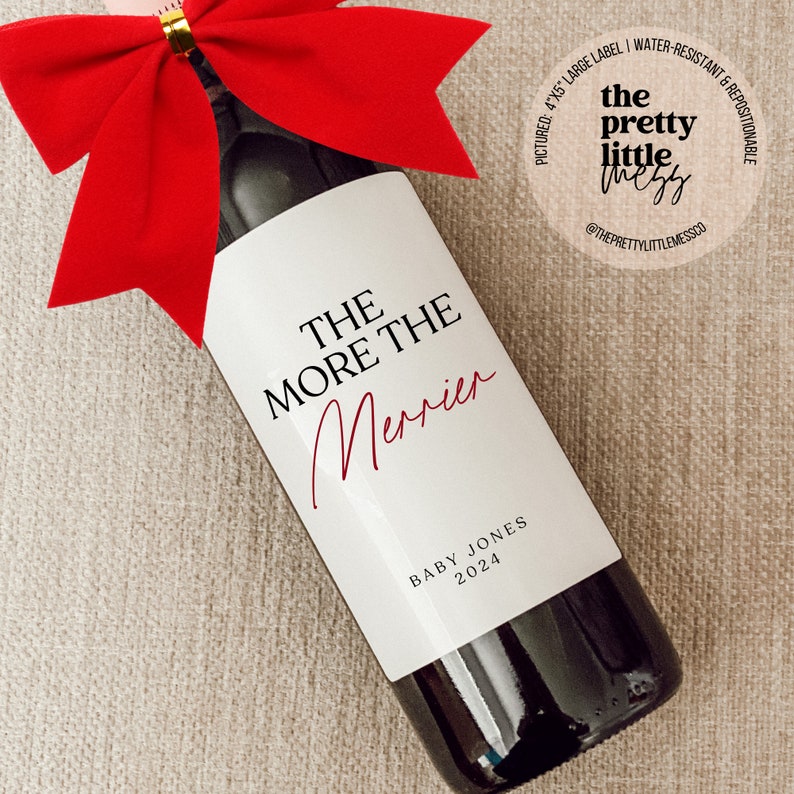 May include: A white wine bottle label with the text "THE MORE THE MERRIER" in black and red lettering. The label also includes the text "BABY JONES 2024" in black lettering.