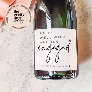 Puede incluir: Etiqueta de botella de champ&aacute;n con fondo blanco y texto negro que dice "Pairs well with getting engaged. Cheers, to forever."