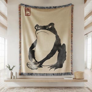 May include: Beige throw blanket featuring a large black illustration of a frog with a bowl. The frog is holding a utensil over the bowl. The blanket has a fringed edge and a red and black square symbol in the upper left corner.