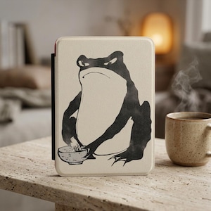 May include: A rectangular e-reader case in a light cream color featuring a black graphic illustration of a frog holding a whisk and a bowl. The case has a textured surface and a thin black spine detail on the left side, presenting a clean design.