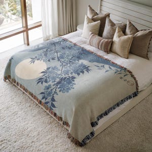 Pode incluir: Uma manta de tecido azul e branco com um design floral e uma lua cheia. A manta está sobre uma cama branca com vários travesseiros.
