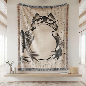 May include: A woven tapestry featuring a black and white illustration of a frog on a beige background with black Japanese characters. The tapestry has a fringe border.