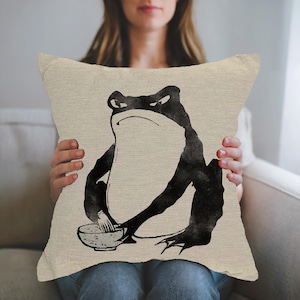 Könnte beinhalten: Beiges quadratisches Kissen mit einer schwarzen Tuscheillustration einer Frosch, der eine Schüssel und ein Utensil hält. Der Frosch hat einen strengen Ausdruck. Das Kissen wird von einer Person gehalten.