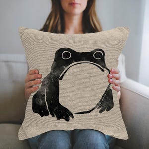 May include: Beige square throw pillow featuring a large black frog illustration. The frog design is a simple, stylized drawing with a minimalist aesthetic. The pillow has a textured appearance.