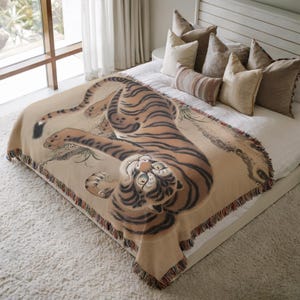 Könnte beinhalten: Eine beige Decke mit einer detaillierten Illustration eines Tigers mit orangefarbenen und schwarzen Streifen. Der Tiger liegt mit nach oben gerichtetem Kopf. Die Decke hat einen Fransenrand und wird auf einem Bett mit Kissen präsentiert.