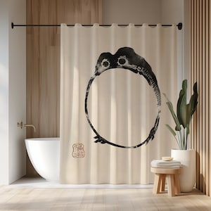 May include: Cream-coloured shower curtain with a large black ink illustration of a circular design with a bird-like head. A red stamp is in the lower left corner. The curtain hangs in front of a white bathtub and wooden accents.
