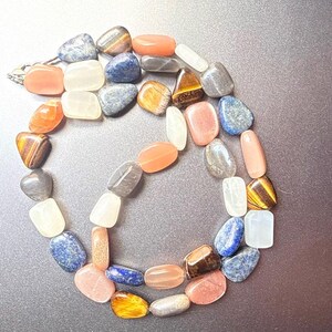 May include: A necklace composed of various polished gemstones in shades of orange, blue, white, gray, and brown. The stones are strung together, forming a circular shape. The necklace is displayed on a dark surface.