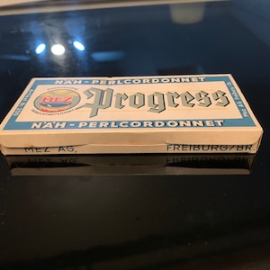 May include: A blue and white box with the text "NAH - PERLCORDONNET" and "Progress" in a stylized font. The box also has the text "MEZ AG, FREIBURG/BR."