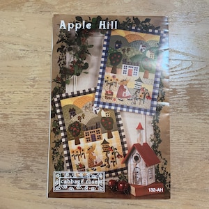 May include: A quilt pattern booklet titled "Apple Hill" featuring two colorful quilt designs with houses, trees, and a woman in a dress. The booklet is from Cabbage Rose, and the pattern number is 132-AH.