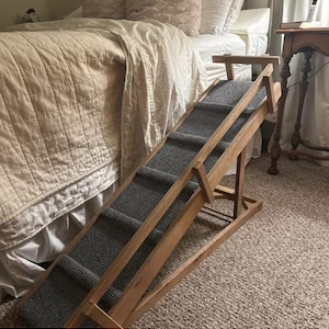 May include: A wooden pet ramp with a gray carpeted surface and side rails. The ramp is positioned at an incline, featuring a sturdy frame with a support structure underneath to maintain its angle, designed to assist pets in reaching elevated surfaces.