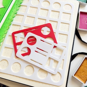 May include: A large white stencil with circular and square cutouts, a smaller red stencil, and a small white stencil with geometric shapes. A green scalloped ruler, watercolour pans in pink and gold, and a paintbrush are also visible.