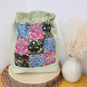 May include: A green drawstring bag with a patchwork design. The patchwork is made of different floral patterns in shades of pink, red, blue, and green. The bag is sitting on a wooden surface.