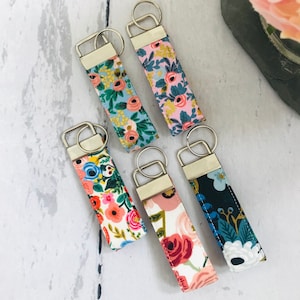 May include: Five colourful floral patterned keychains with silver key rings. The keychains are made of fabric and have a variety of floral patterns, including pink, blue, and white.