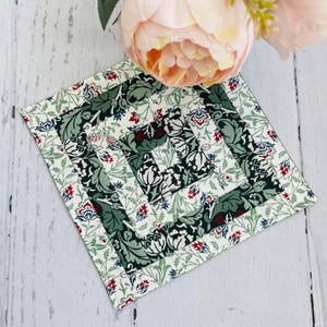 May include: A square fabric coaster with a green and white floral pattern. The coaster has a red and white border.