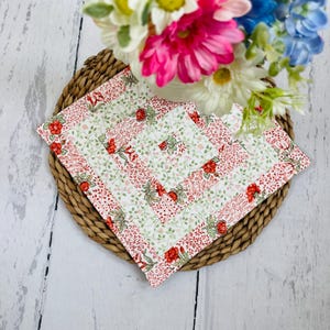 May include: Four square fabric coasters with a floral pattern in red and green on a white background. The coasters are arranged on a woven wicker placemat, with a bouquet of flowers in the background.