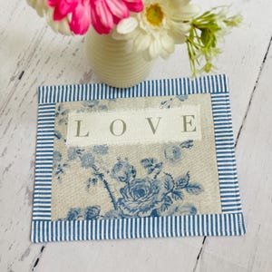 May include: Square fabric coaster featuring a blue floral design and the word "LOVE". It has a blue and white striped border. A vase of flowers is visible in the background. The coaster is likely used for placing drinks.