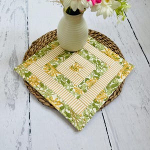 May include: A square, quilted trivet with a floral and striped pattern. The trivet has a cream background with yellow stripes and green floral accents. It rests on a woven, round placemat, with a cream vase holding flowers.