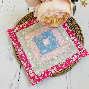 May include: A square, quilted coaster with a floral pattern in shades of pink, grey, and blue. The coaster has a bright pink border with white floral accents. The coaster is placed on a woven, circular mat.