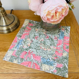 May include: A square, quilted coaster featuring a floral design in shades of pink, green and blue. The coaster has a white background and a decorative border.