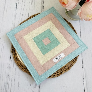 May include: A square quilted hot pad with a layered design in shades of blue, pink, and cream. The hot pad features a smaller square in the centre with a "LIBERTY. FABRICS" label.