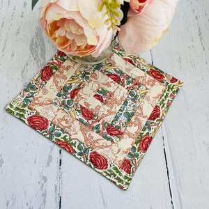May include: A square quilted coaster featuring a floral design in shades of red, green and cream. The coaster has a white background with a repeating pattern of stylised flowers and leaves.