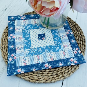 May include: A square fabric quilt block featuring a log cabin pattern with concentric borders of blue and white floral, striped, and polka dot prints. The centre square is dark blue with a small floral motif, surrounded by light blue polka dots.