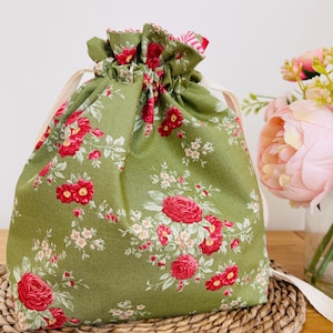 May include: A green drawstring bag featuring a floral design of red roses and white blossoms. The bag has a gathered top and cream drawstrings. It is placed on a woven mat, with a floral arrangement in the background.