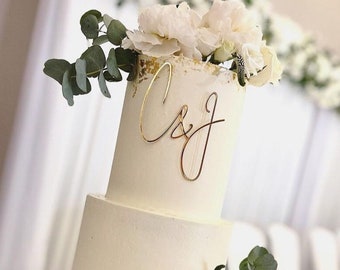 May include: A three-tiered white wedding cake decorated with gold leaf and white flowers. The top tier has a gold topper with the initials "C&J".