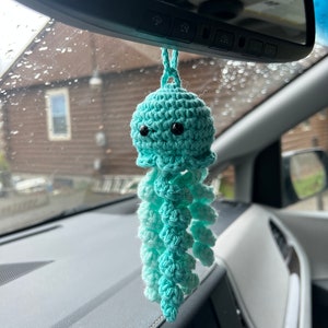 May include: A teal blue crocheted jellyfish car charm hanging from the rearview mirror of a car. The jellyfish has black eyes and a long, flowing tail.