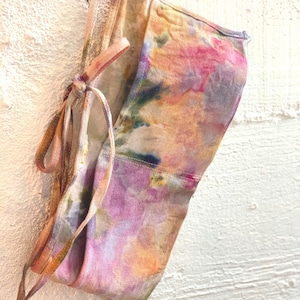 May include: A tie-dye fabric face mask in shades of pink, purple, orange, and green. The mask has a tie closure and is made of a soft, lightweight material. The mask is hanging against a white textured wall.