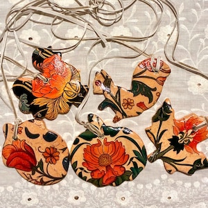 May include: Five flat, shaped ornaments made of a light tan material, each featuring hand-painted floral and botanical designs in vibrant orange, red, green, and black. Each ornament is suspended by a thin, light-colored cord for hanging.