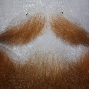May include: A close-up of a white mannequin head with a red-brown beard and mustache. The beard and mustache are made of hair fibers and are styled in a classic look.
