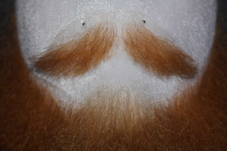 May include: A close-up of a white mannequin head with a red-brown beard and mustache. The beard and mustache are made of hair fibers and are styled in a classic look.
